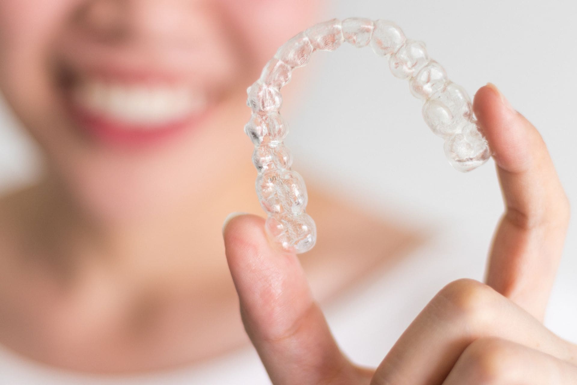A close-up shot of a person holding up a clear plastic Invisalign tray in their hand, with their blurred, smiling face visible in the background.